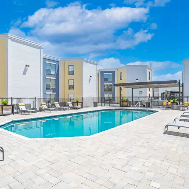 A sunny outdoor pool area with lounge chairs and umbrellas, surrounded by modern apartment buildings.