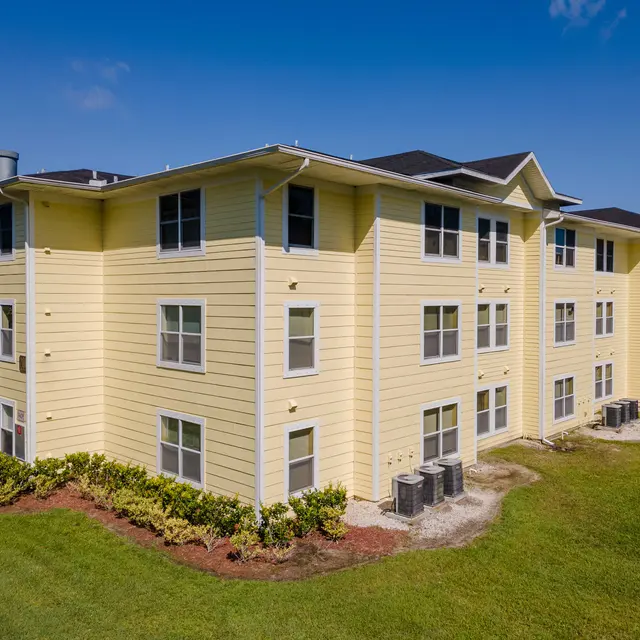 A yellow three-story apartment building surrounded by greenery and open grass.