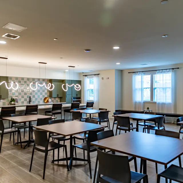 A modern dining area featuring multiple tables and chairs, with a kitchen bar in the background and contemporary lighting fixtures.