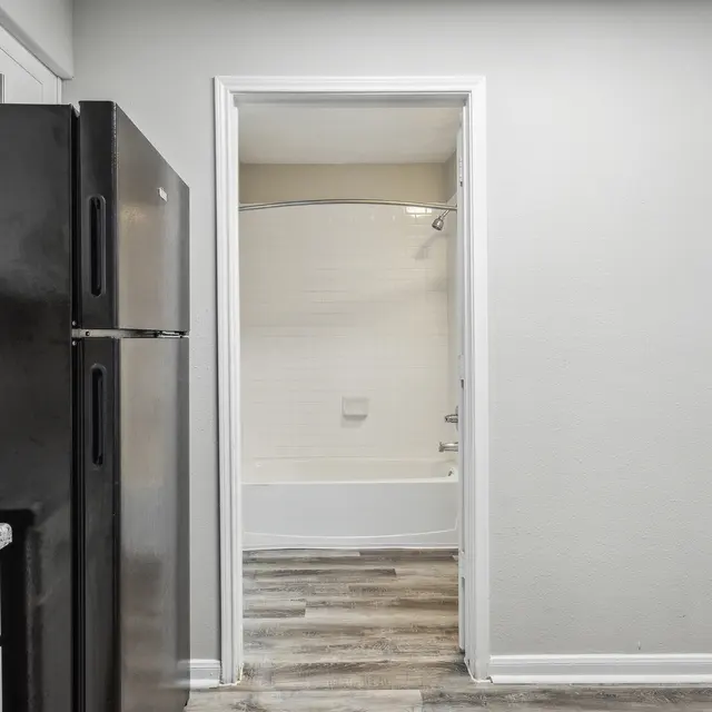 View of a kitchen with black appliances, leading to a bathroom doorway featuring a tub and shower. Neutral wall colors.