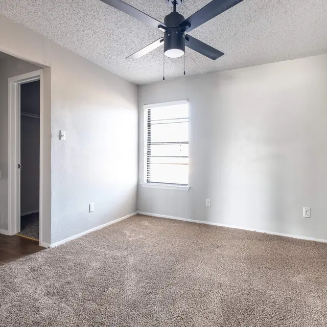 An empty room featuring beige carpet, a ceiling fan, and a window with blinds. There is a doorway leading to another room visible and a vanity with a mirror on one side.