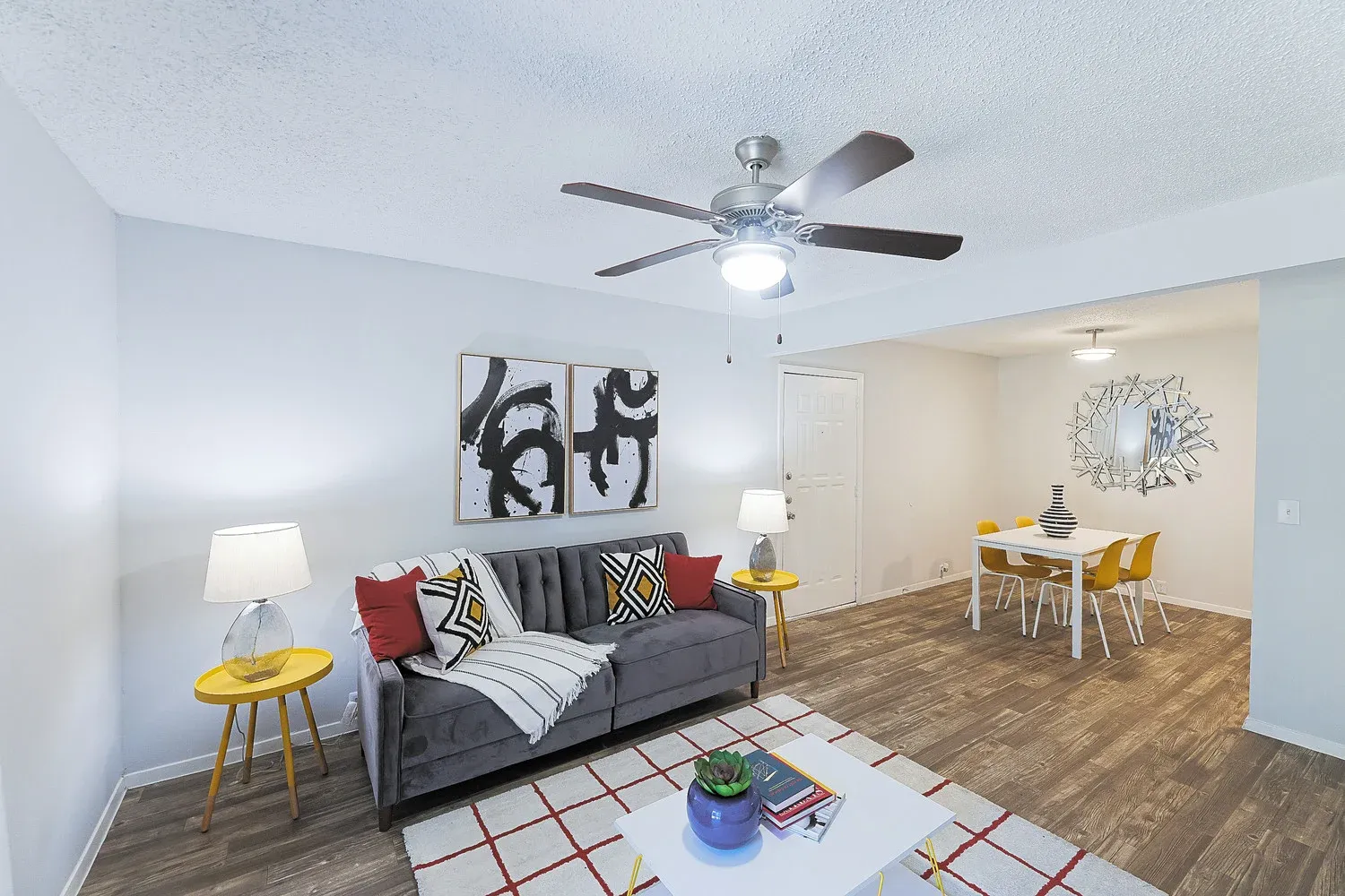 A modern living room featuring a gray sofa with colorful throw pillows, two yellow accent tables, and a white coffee table with decorative items. On the walls are abstract art pieces. The room has a ceiling fan and is connected to a dining area with a table and chairs.