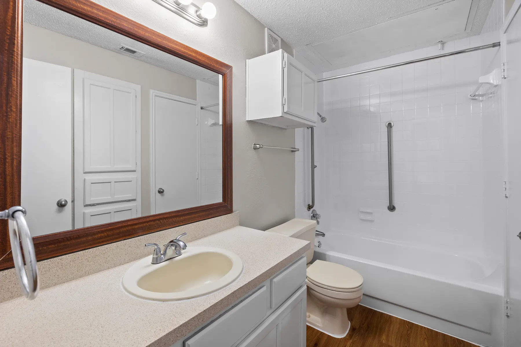 A clean and well-lit bathroom featuring a bathtub, toilet, and sink with a countertop. A large mirror above the sink reflects the space, and storage cabinets are visible on the wall. The flooring appears to be wood-like.