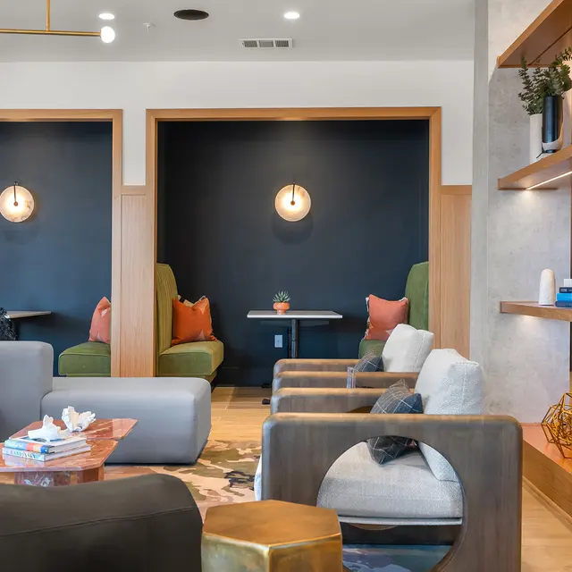 A modern lounge interior featuring cozy seating arrangements with green chairs, a small wooden table, and decorative elements. There are books on the coffee table and wall shelves with plants and sculptures.