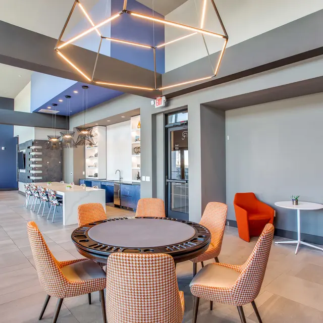 A spacious modern lobby area featuring a round table with stylish chairs, colorful accent chairs on the sides, and a well-lit open space with a kitchen in the background.