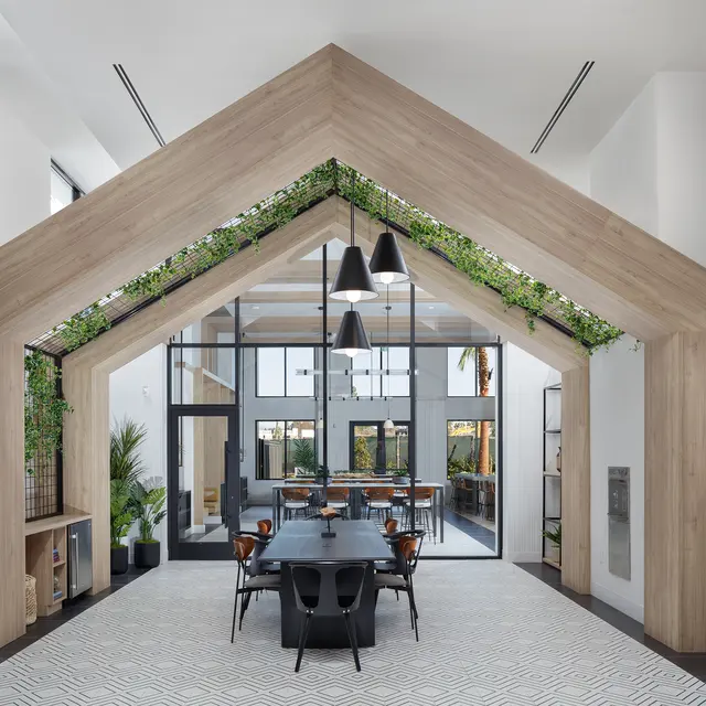 A spacious dining area featuring a distinctive wooden framed ceiling with greenery, black pendant lights, and a long black dining table surrounded by stylish chairs.