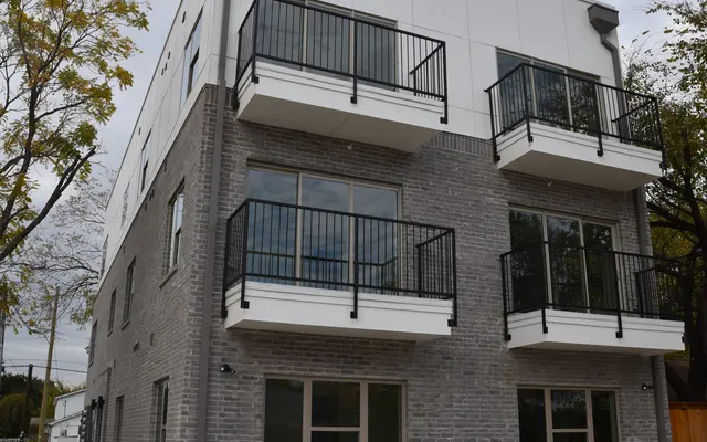 Three-story building with gray brick and white siding, featuring balconies with black railings and an empty parking area in front.