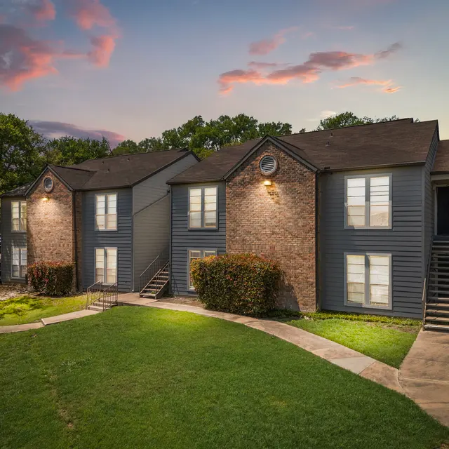 A residential apartment complex with brick and blue siding, featuring grassy areas and a walkway, during dusk with a colorful sky in the background.