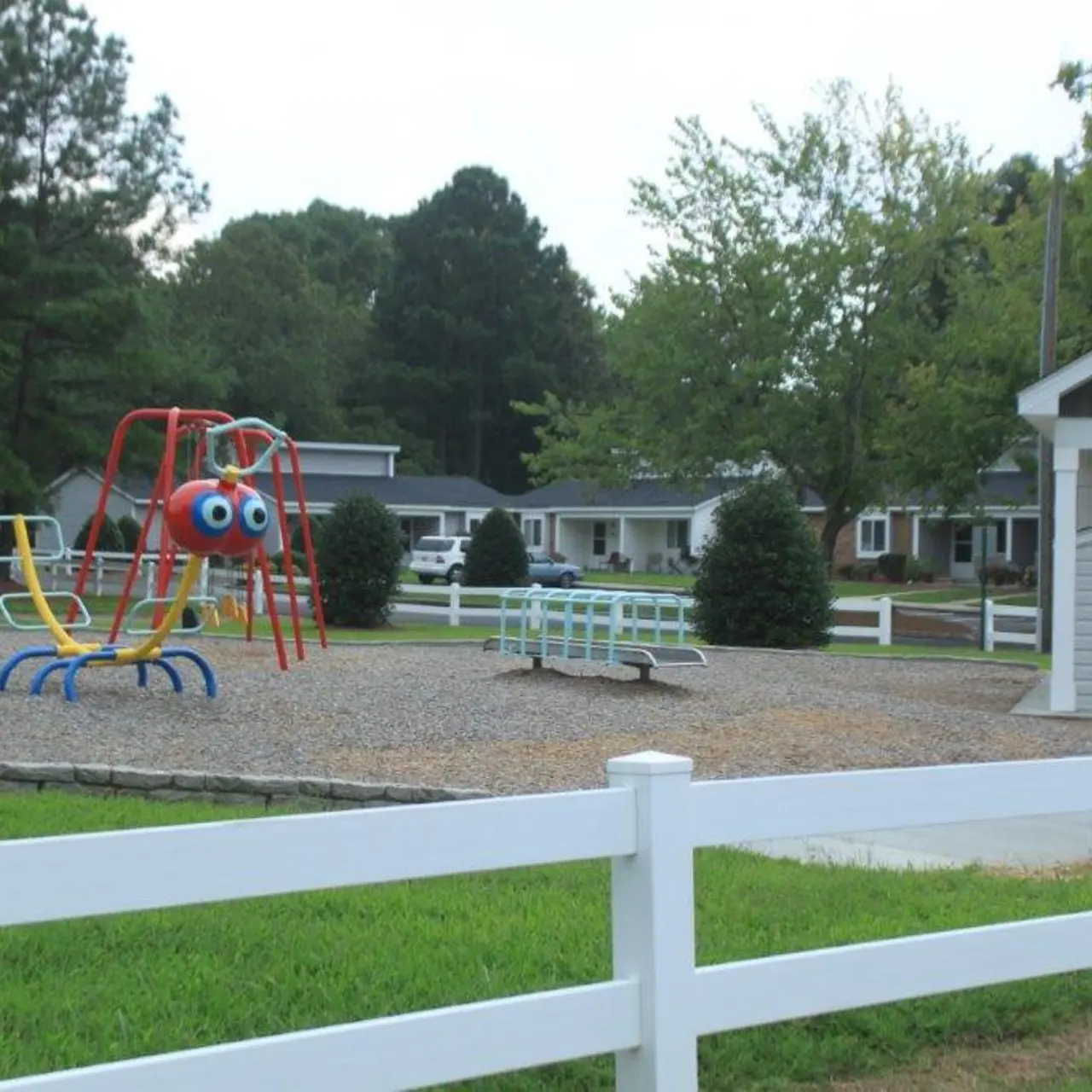 Colorful playground equipment in a community park, featuring a spider-shaped climbing frame and benches, surrounded by grass and trees.