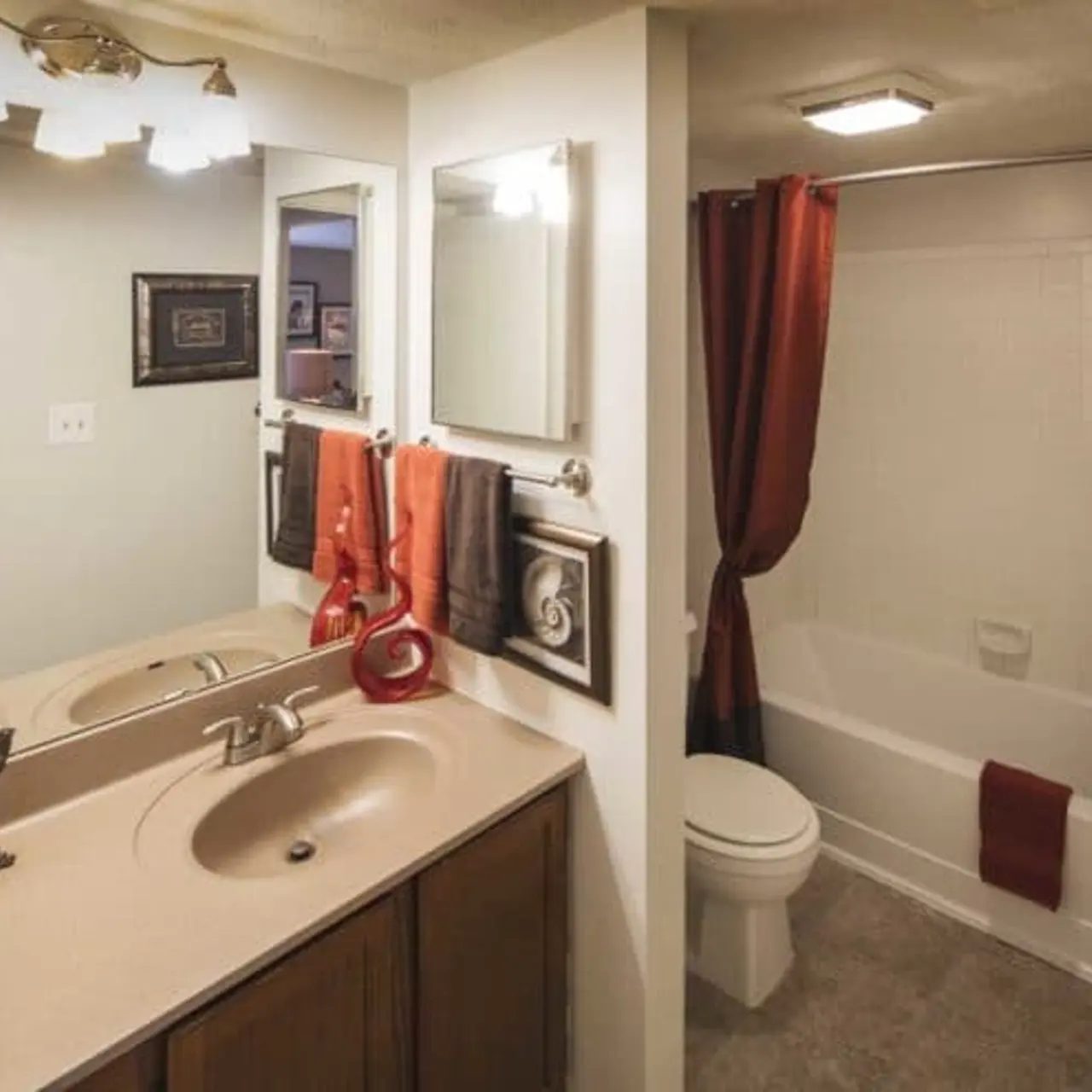 Modern bathroom with double sinks, red curtains and towels, a white bathtub, and a framed mirror above the sink under a light fixture.