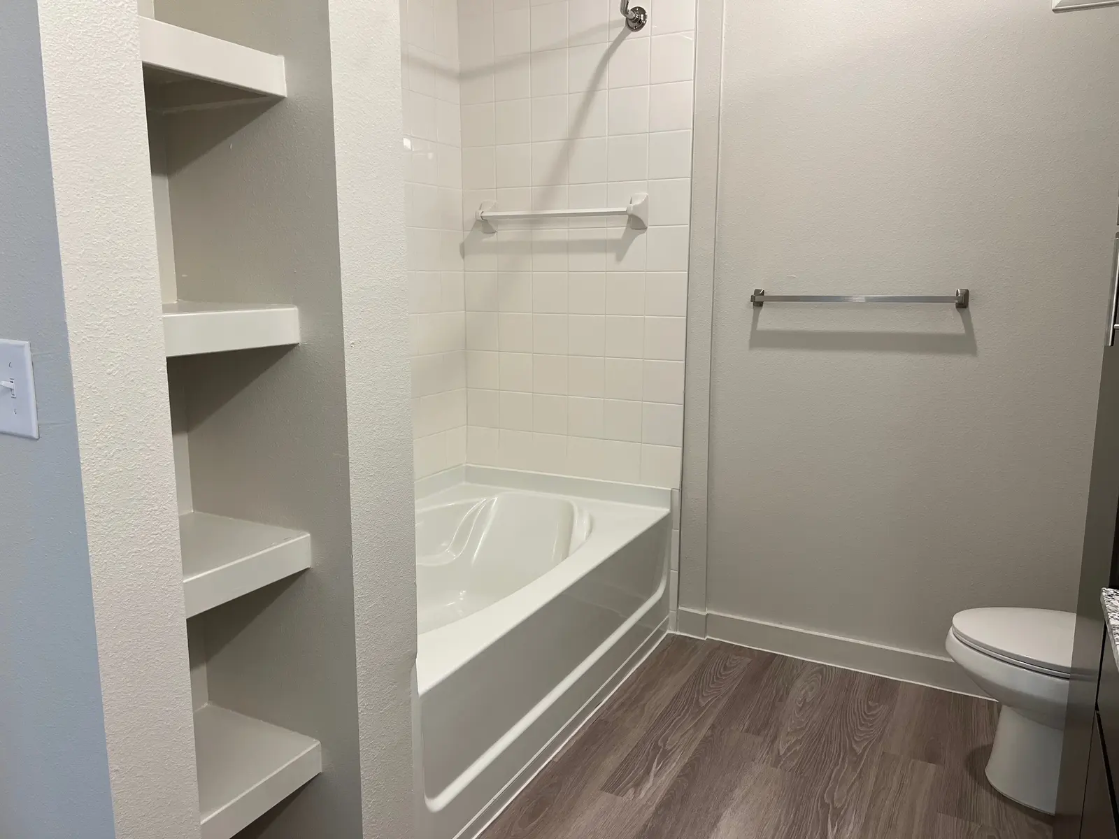 A modern bathroom featuring a white bathtub, shower, and shelving. The walls are painted light gray and the flooring is dark brown laminate.