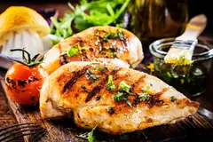 Two large chicken breasts with grill marks lay on a cutting board. Grilled tomatoes, lettuce, and a bowl of oil surround the chicken. 