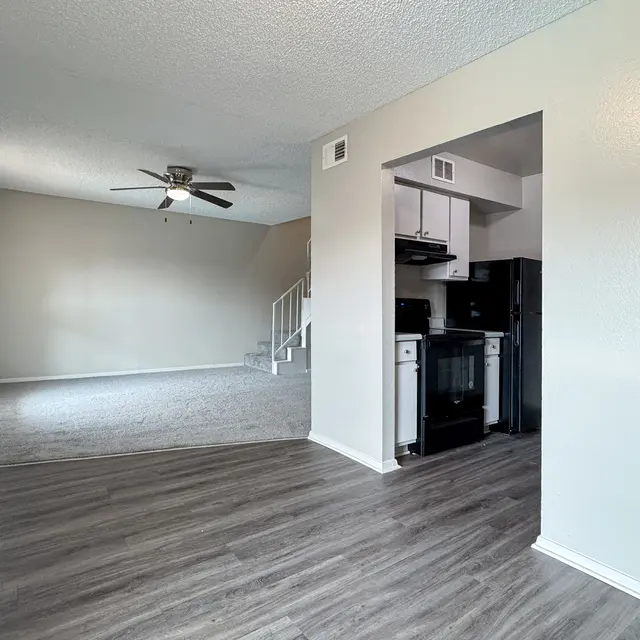Interior view of a modern apartment featuring a living area with a ceiling fan, carpeted flooring, and an open kitchen design with black appliances.