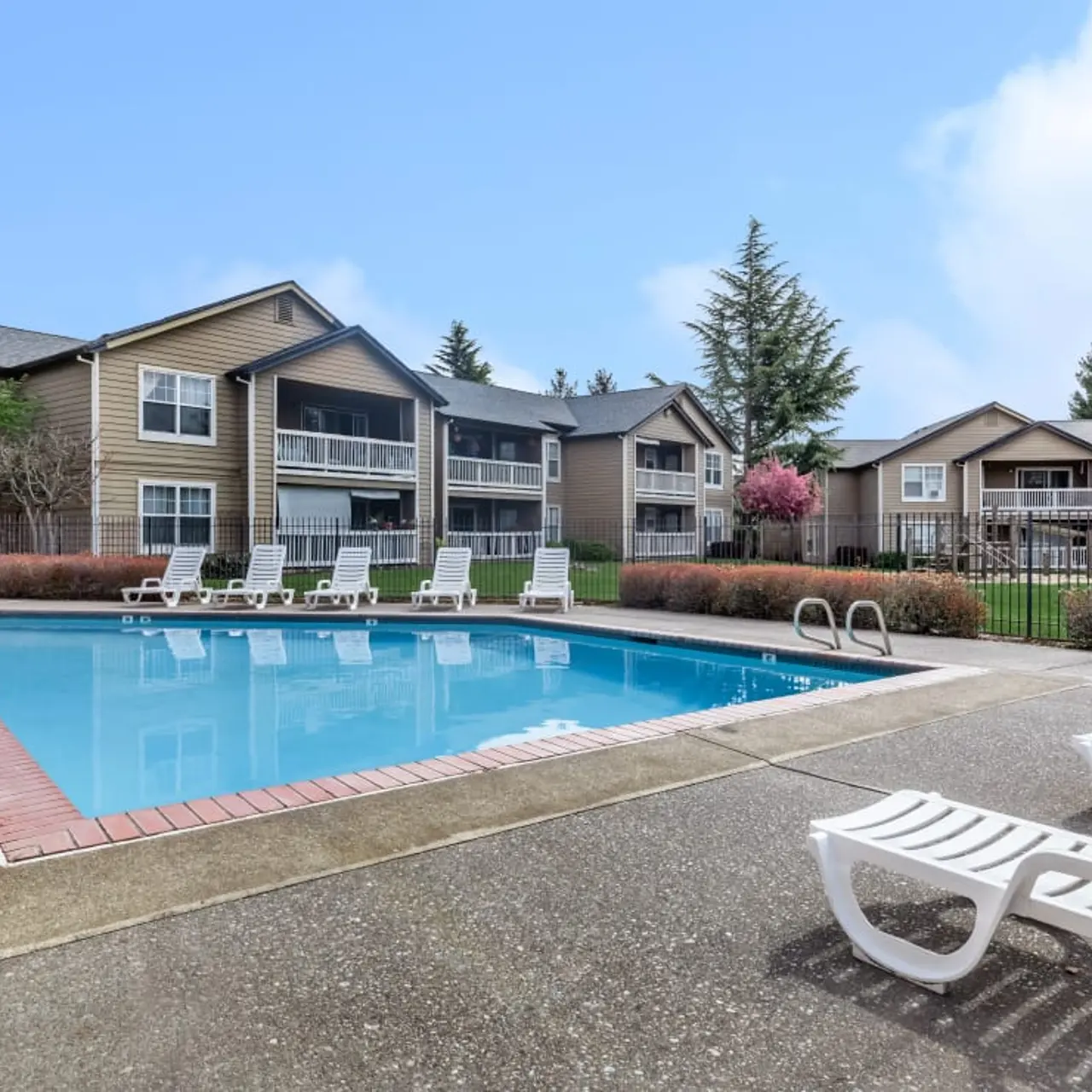 A view of a swimming pool surrounded by lounge chairs, with several apartment buildings in the background and greenery around the area.