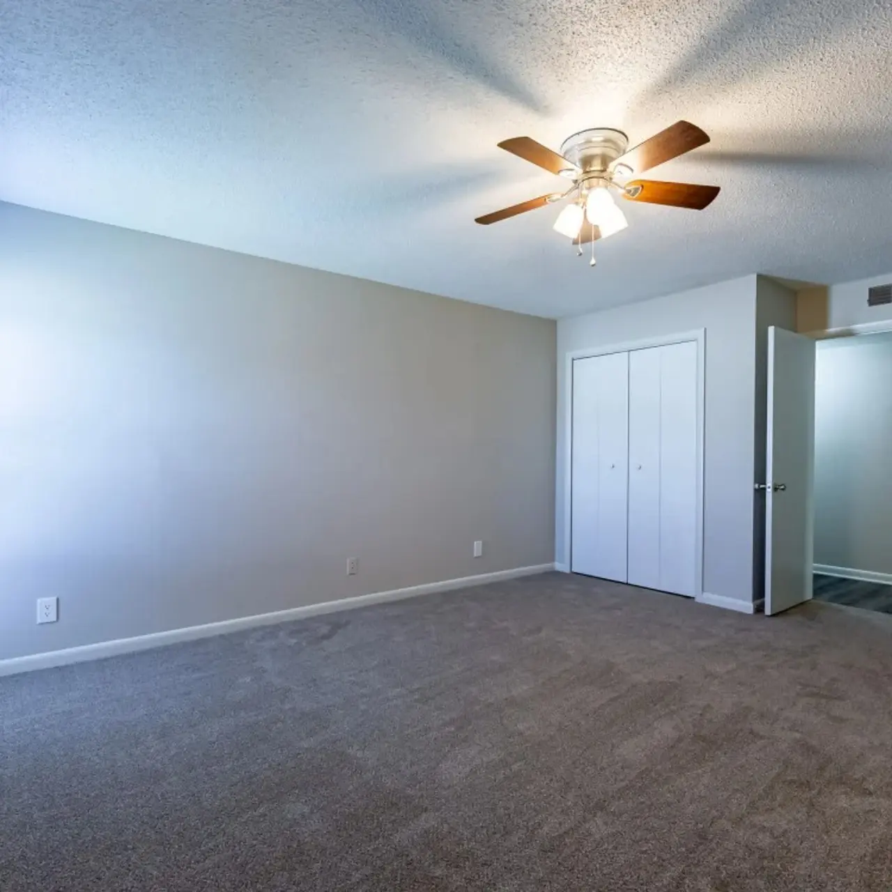 An empty room with light gray walls and carpet, featuring a ceiling fan and closet doors. Natural light enters through windows on one side.
