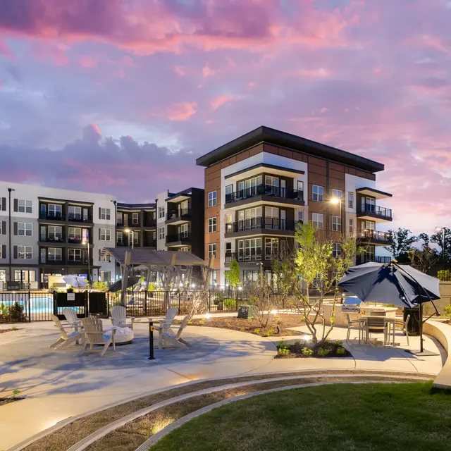 A modern apartment complex with a pool area at dusk, featuring landscaped gardens and outdoor seating.