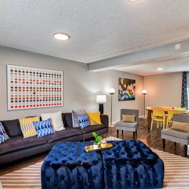 A modern living room featuring a dark sectional sofa with bright throw pillows, a stylish blue ottoman, and artistic wall decor. A dining area with yellow chairs is visible in the background.