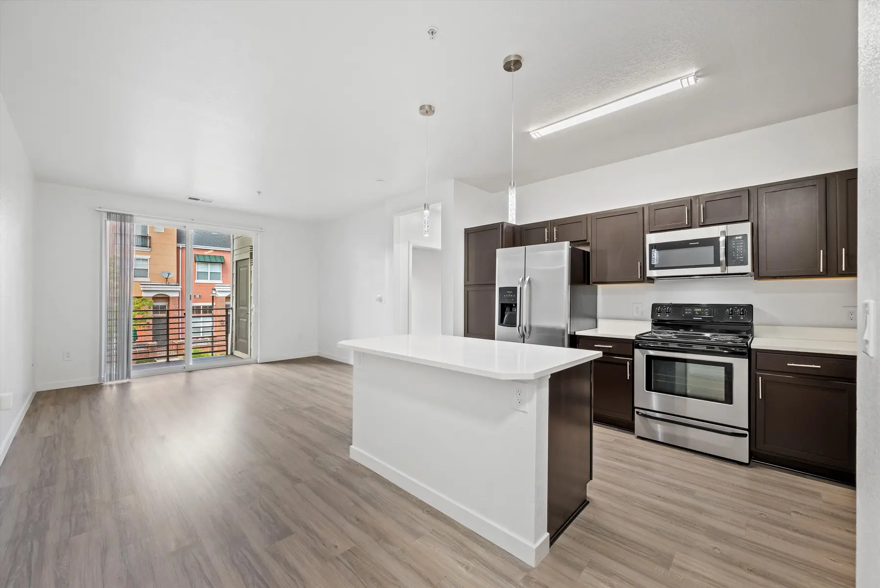 Interior view of a modern kitchen and living room with an open layout, featuring a kitchen island, dark wood cabinetry, and large windows overlooking a balcony.