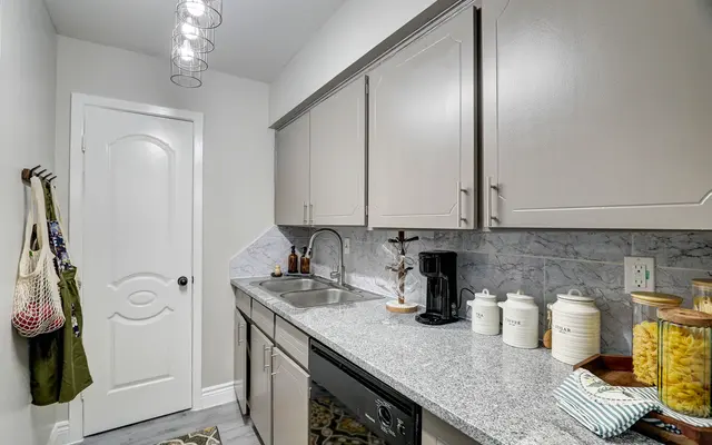 A modern kitchen with gray cabinets and a granite countertop, featuring a sink, coffee maker, and decorative jars. The walls are a light color, and there is a door leading to another room.