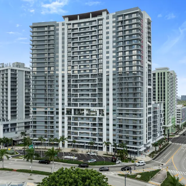 A view of a modern high-rise building complex with multiple towers. The architecture features glass balconies and a sleek design, surrounded by palm trees and a city street.