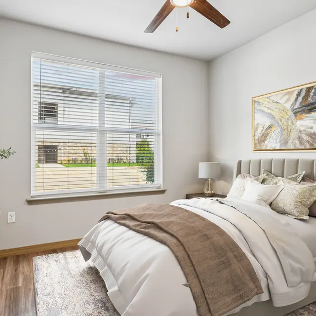 A cozy bedroom featuring a bed with a light gray headboard, white bedding, a decorative throw, and pillows. A nightstand with a lamp is next to the bed. A window with blinds allows natural light to flood the room. A piece of abstract art hangs on the wall, and green plants are present on the nightstand and dresser.