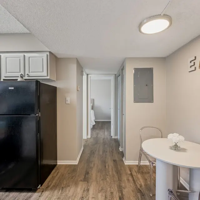 A small, modern kitchen in an apartment featuring a black refrigerator, gray cabinets, and a white dining table with two chairs.