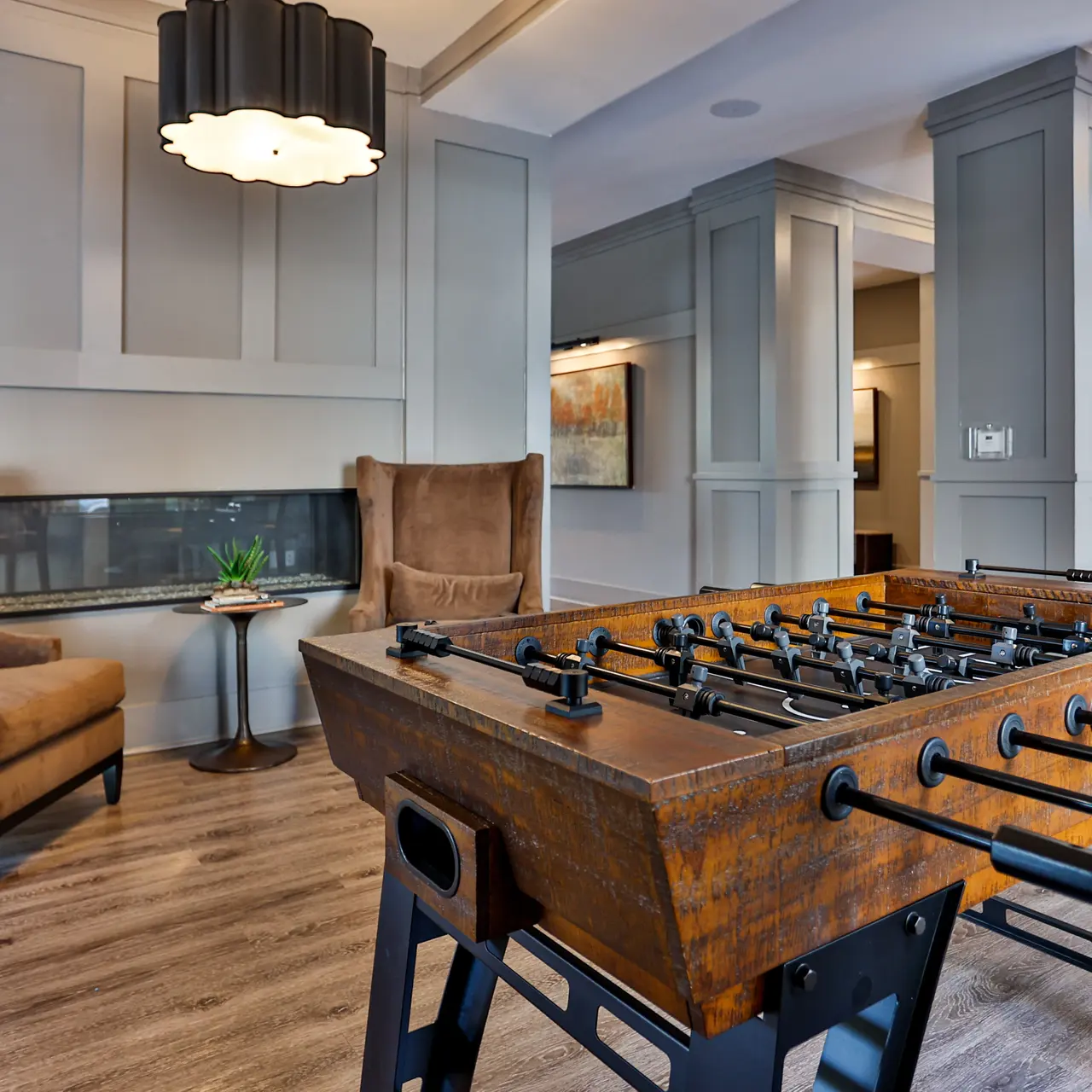 A modern lounge area featuring a foosball table and two stylish armchairs, with a decorative table and a plant in between. The space is well-lit and stylishly designed.