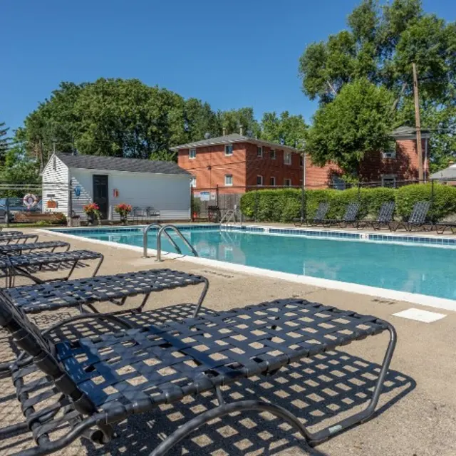 An outdoor swimming pool surrounded by lounge chairs on a sunny day. Lush green trees and residential buildings are visible in the background.