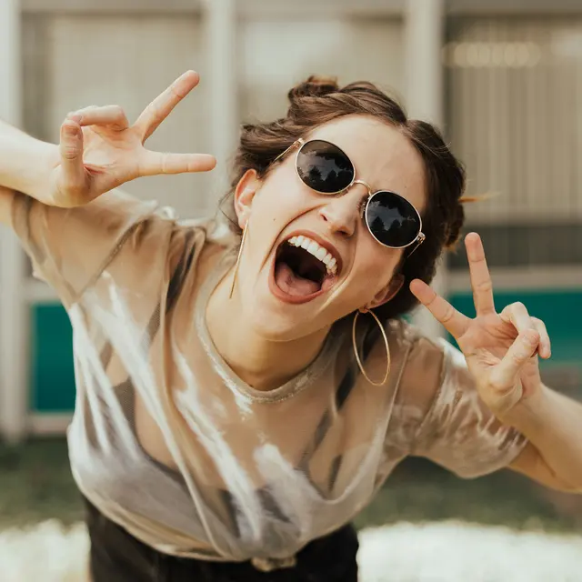 A young woman in sunglasses making a playful peace sign gesture with her hands. She has a bright smile and is wearing a casual outfit with a light top and shorts, standing in an outdoor setting.