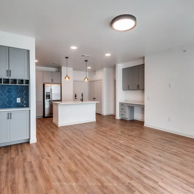 A modern apartment interior featuring an open layout with a kitchen and living room. The kitchen has grey cabinets and a white island, with pendant lights above. There is a blue accent wall and wooden floors throughout the space.