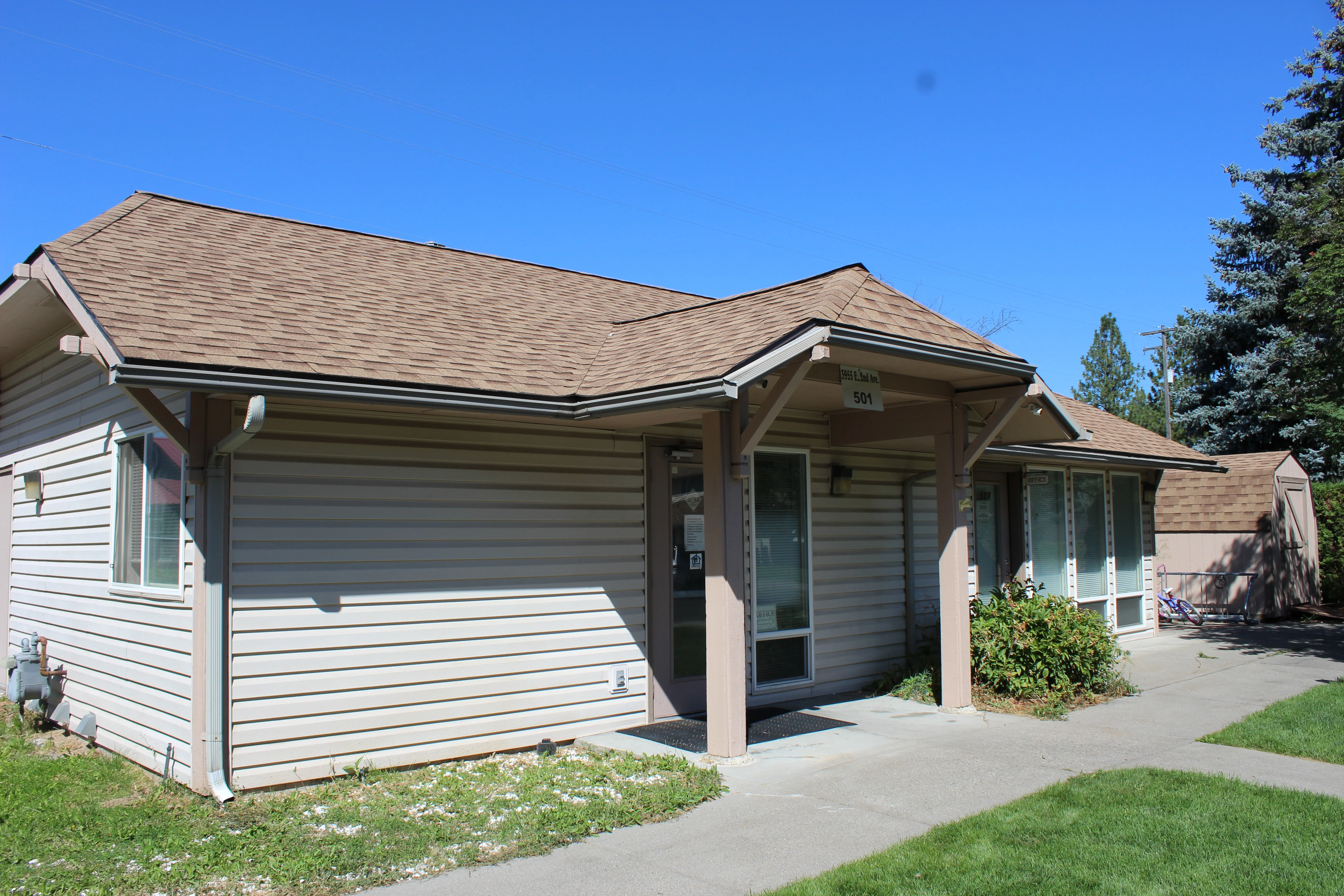 An exterior view of a single-story residential bungalow with a tan siding and a brown roof, featuring a small garden area in front and a clear blue sky overhead.