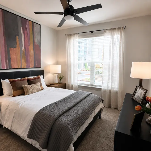 A modern bedroom featuring a queen-sized bed with a gray blanket and decorative pillows. There are two bedside tables with lamps, a large window with sheer curtains, and a colorful abstract painting on the wall. A dresser is adorned with a vase of tulips and a picture frame.
