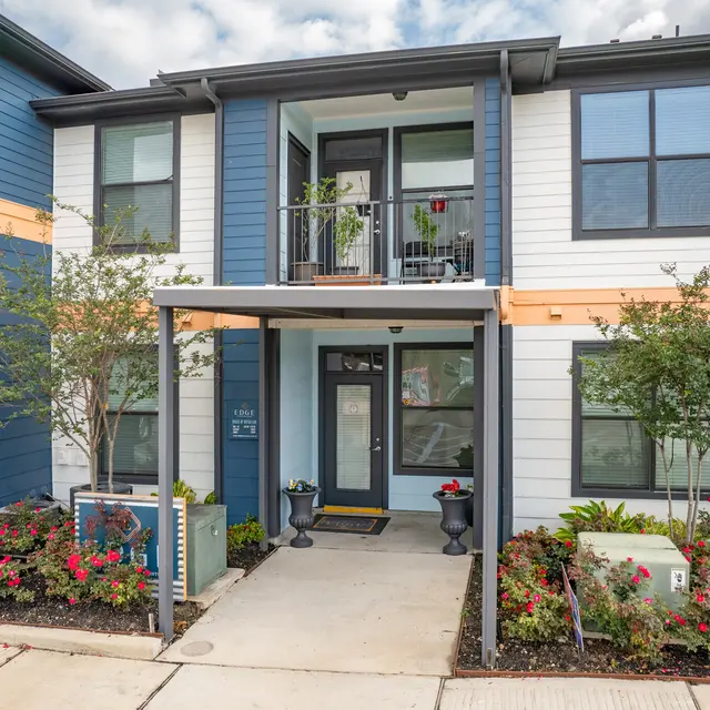Exterior view of a modern multi-unit apartment building with blue and orange accents, featuring a landscaped entrance and porch.