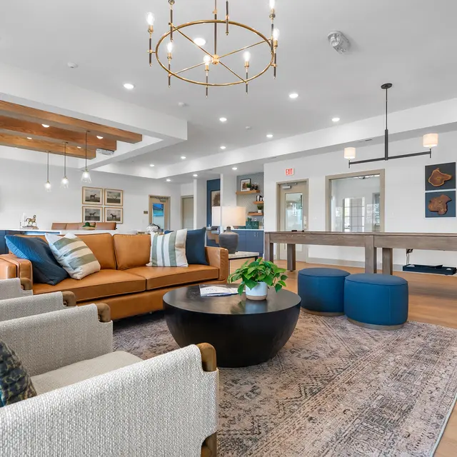 A modern living area featuring a brown leather sofa, light-colored armchairs, a round coffee table, and a large area rug. The space is well-lit with multiple light fixtures and has decorative artwork on the walls.