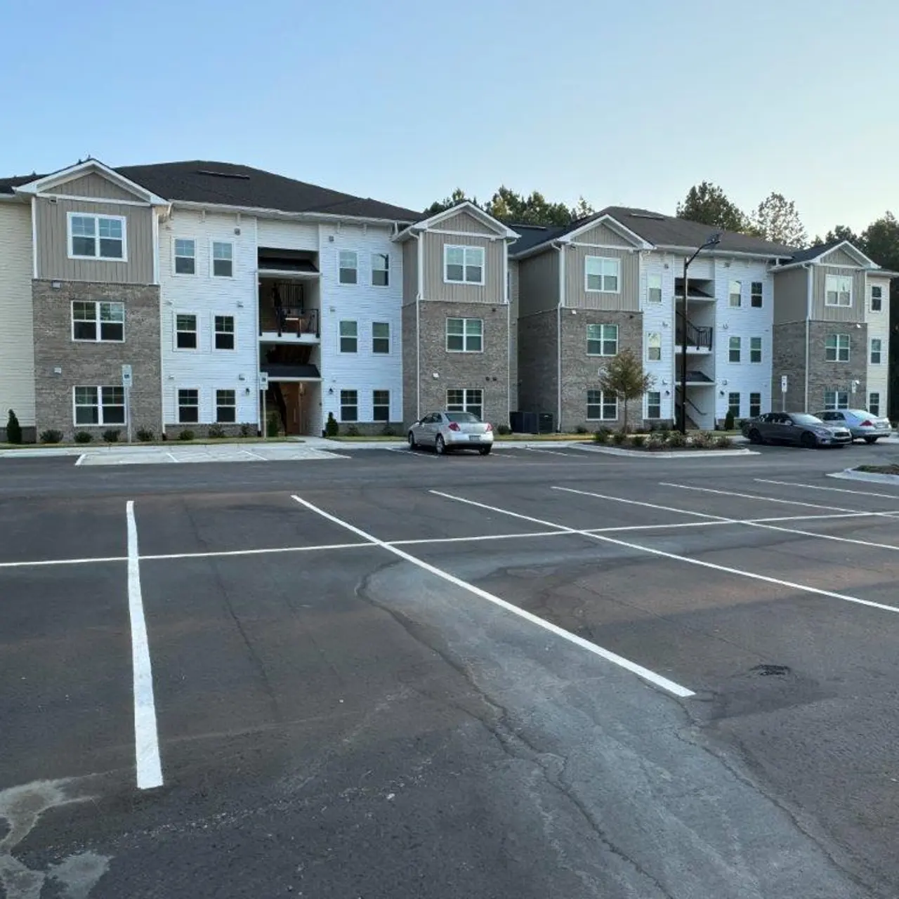 A modern apartment complex with three stories and a landscaped parking area in front. The building features a mix of stone and siding exterior.
