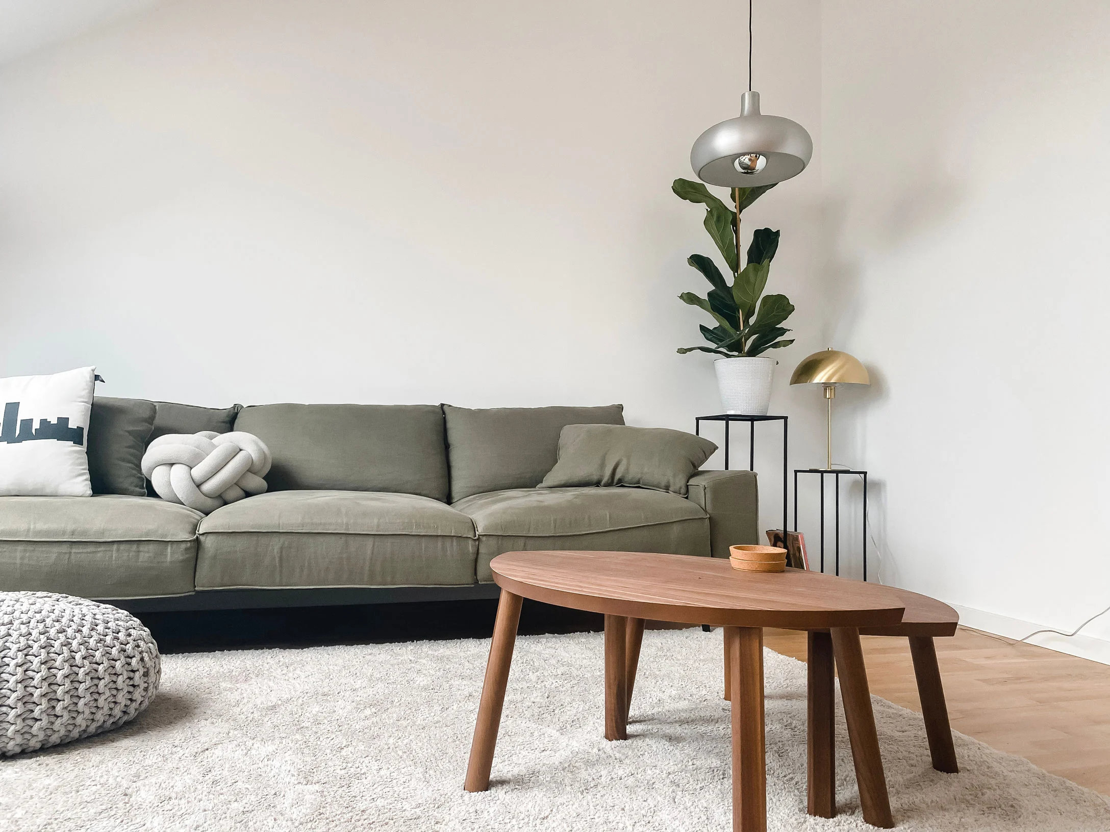 A modern living room featuring a green sofa, a round wooden coffee table, and a cozy grey pouf. There are potted plants and metallic accents visible in the decor.