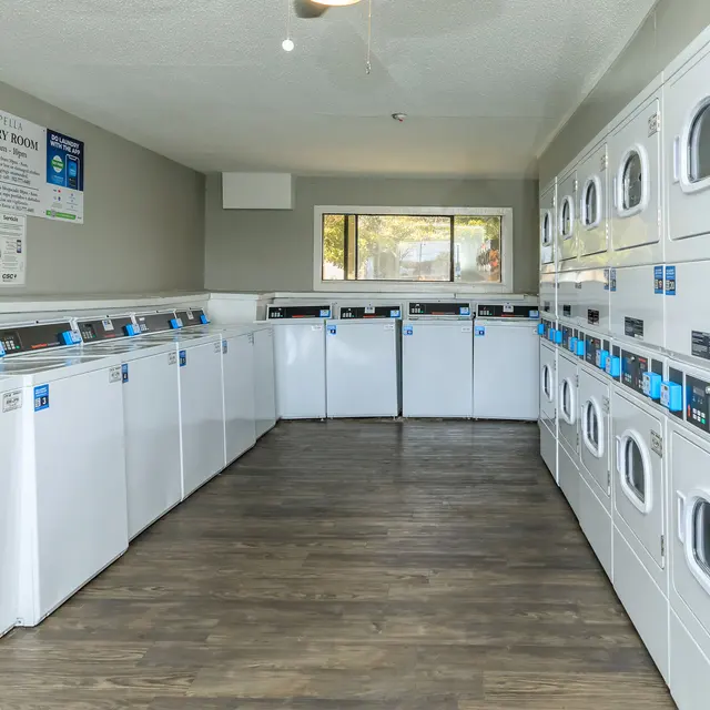 A well-lit laundry room featuring rows of white washing machines and dryers along the walls. The floor is wooden, and there is a large window providing natural light. A sign on the wall displays laundry instructions and information.