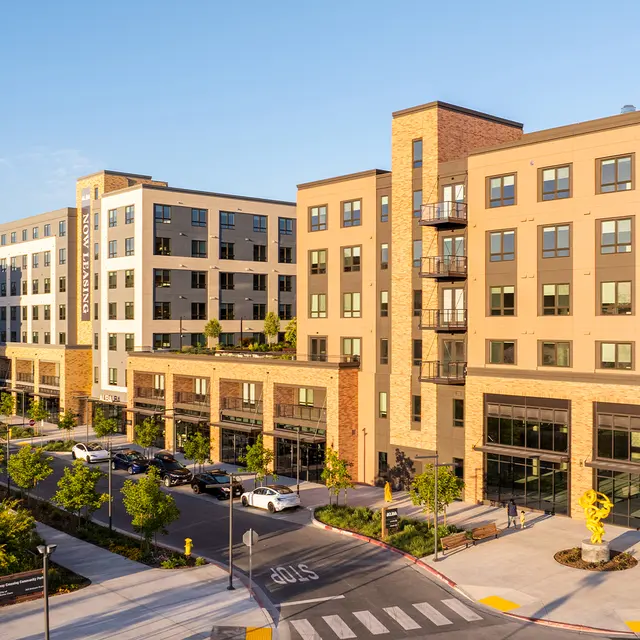 A modern apartment complex with multiple buildings, featuring balconies, large windows, and landscaped surroundings. The foreground has pedestrian pathways and trees.