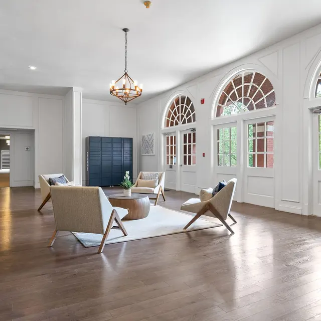 A spacious and modern living room with large windows, hardwood flooring, and light-colored furniture arranged around a round table. Decorative elements include a chandelier and a neutral rug on the floor.