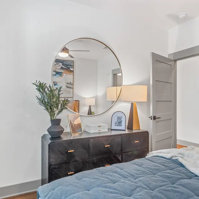 A modern bedroom with a bed featuring a blue quilt, a large round mirror above a dresser, a lamp, and decorative plants.