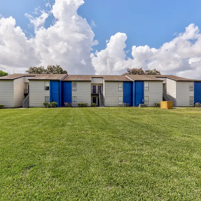Exterior view of a residential apartment complex with blue doors and well-maintained grass in the foreground under a partly cloudy sky.
