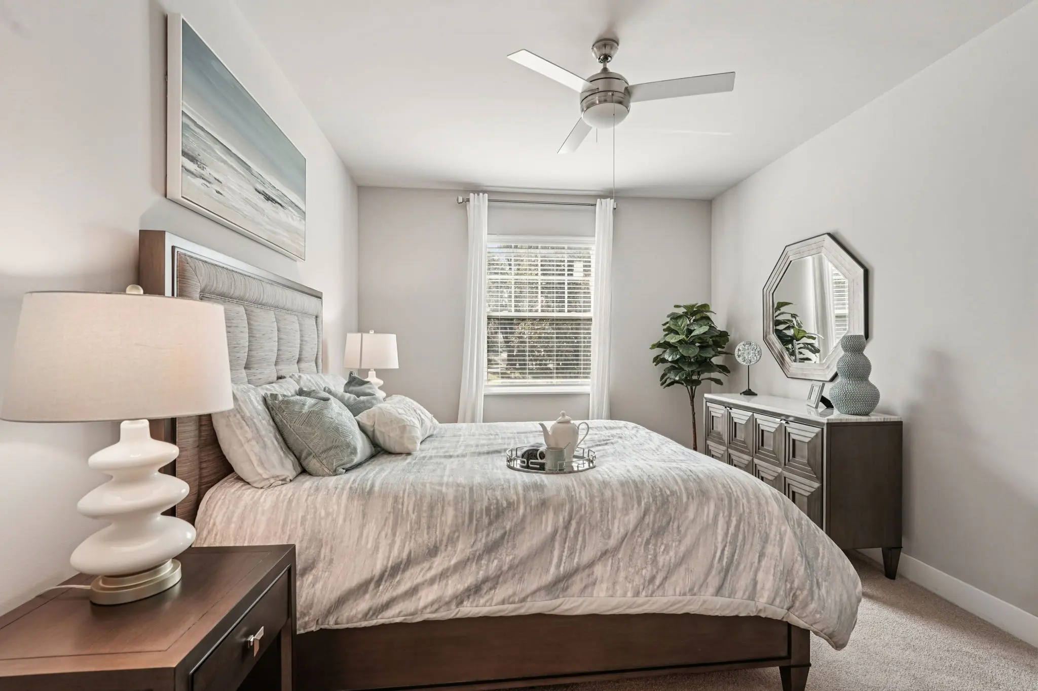 A cozy modern bedroom featuring a queen-sized bed with gray bedding, two bedside lamps, and decorative pillows. A small tray with a teapot and cups sits on the bed. There's a dresser and a plant in the corner, and natural light coming in from a window. The walls are painted white and a piece of art is hung above the bed.