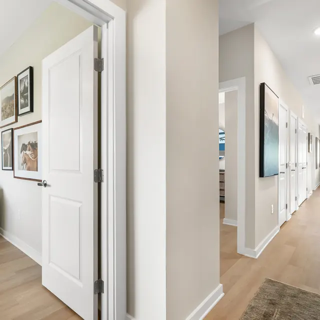 A modern interior hallway featuring light-colored walls and wooden flooring, with a view into a cozy room on the left adorned with framed pictures, and several doors leading to other rooms along the right side.