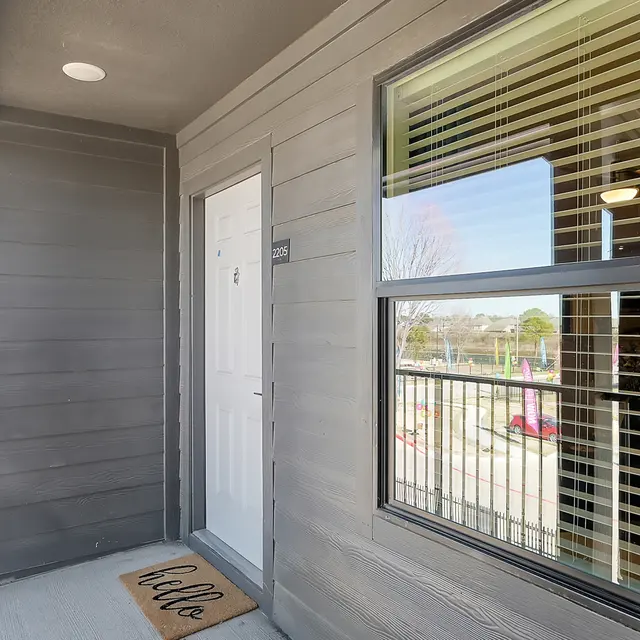 A modern apartment entrance with light gray siding, a white door, and a welcome mat that says 'hello'. Large window with blinds visible adjacent to the door.