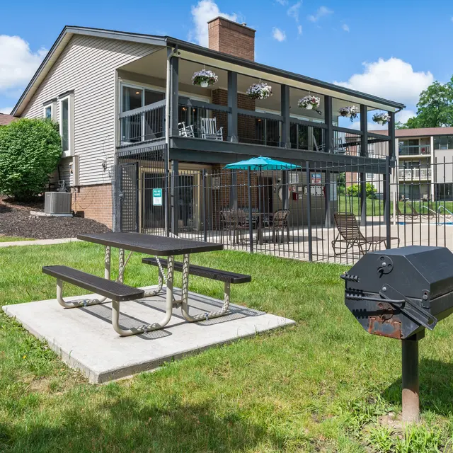 A scenic view of a pool area at an apartment complex featuring a grassy lawn, picnic table, barbecue grill, and a multi-level building with a balcony overlooking the space.