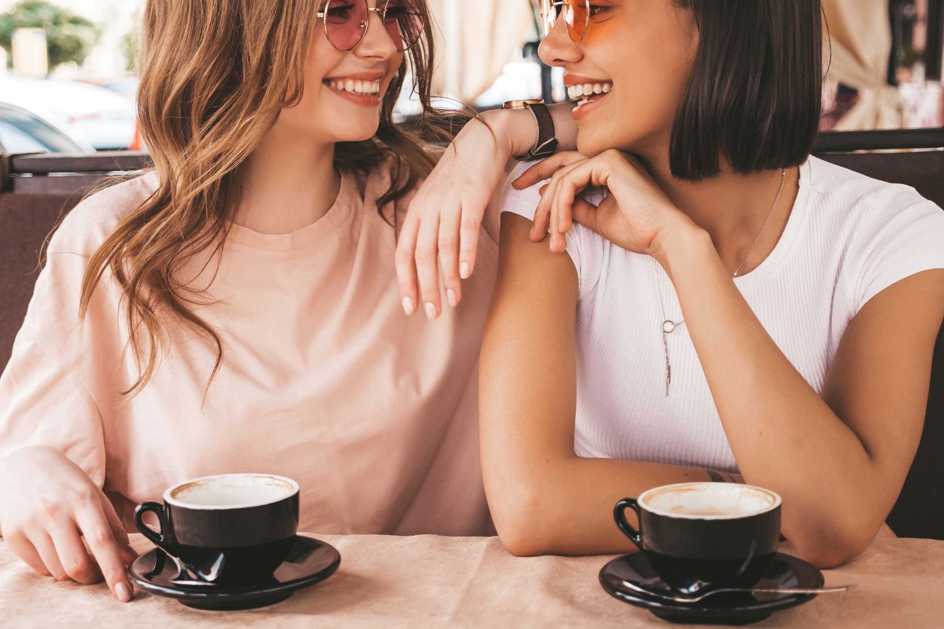 Two women sitting at a café table, smiling and chatting over coffee. Both are wearing sunglasses and casual clothing.