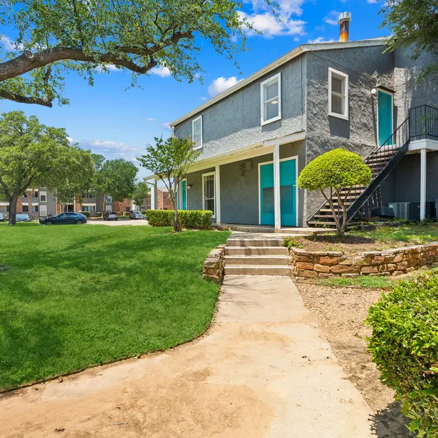 A green lawn with a walking path leading to a two-story gray apartment building. The building has a staircase and several grassy areas with trees and shrubs around it.