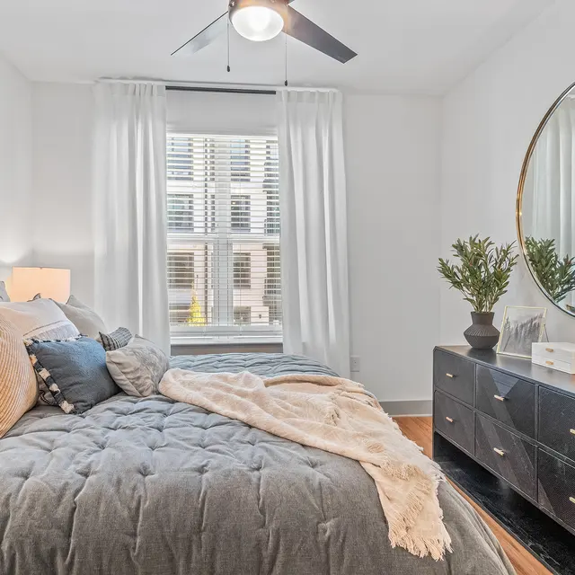 A modern bedroom featuring a bed with gray and orange pillows, a cozy throw blanket, a large round mirror, and a dresser with a lamp and plant.