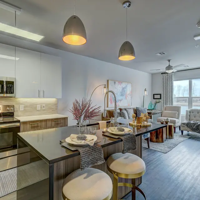 A stylish modern apartment interior featuring a kitchen with stainless steel appliances, a dining area with bar stools, and a cozy living space with a sofa and chairs. Large windows allow natural light to illuminate the space.