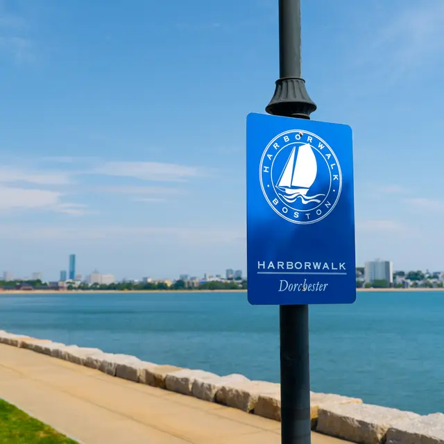 A blue sign displaying 'Harborwalk Boston Dorchester' next to a serene waterfront with a clear sky and city skyline in the background.